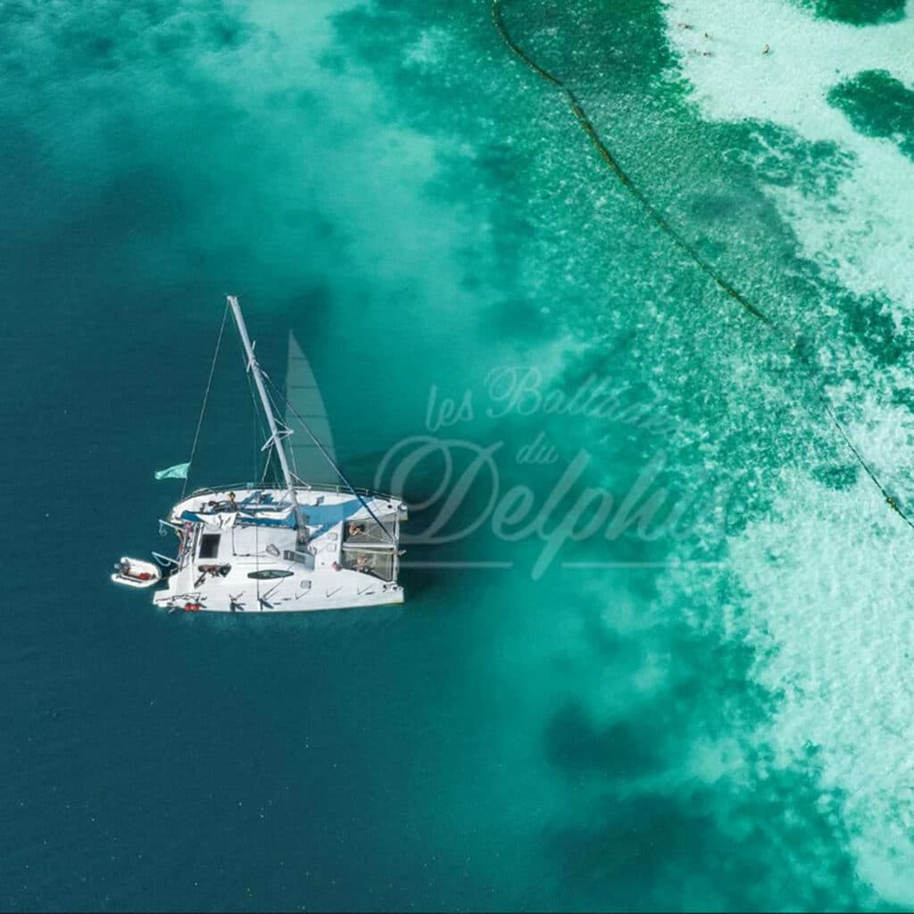 Aerial view of a white catamaran sailboat in bright turquoise sea, with a long wake along a reef on the right.