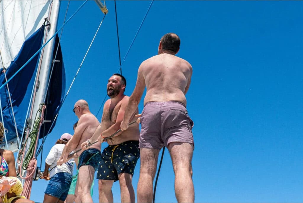 Group of shirtless men standing on a sailboat deck, laughing as they haul lines against a clear blue sky.