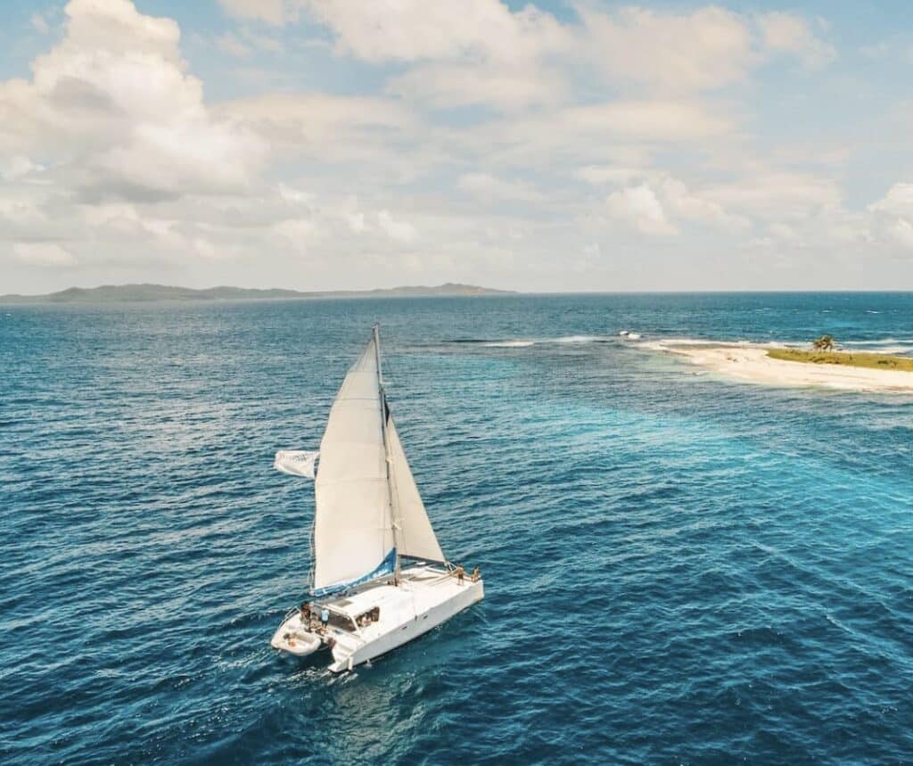 White catamaran sailboat sailing in bright blue ocean toward a sandy island on a sunny day.