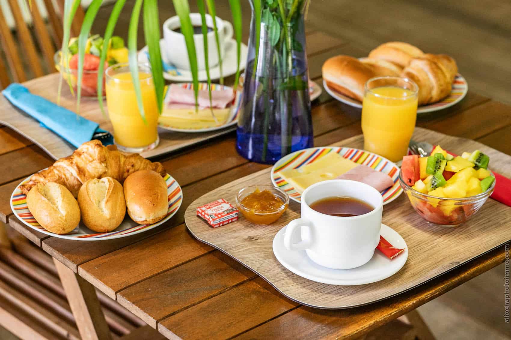 Breakfast spread on a wooden table: croissants, bread rolls, fruit salad, cheese, ham, orange juice, and coffee on striped plates and bowls.