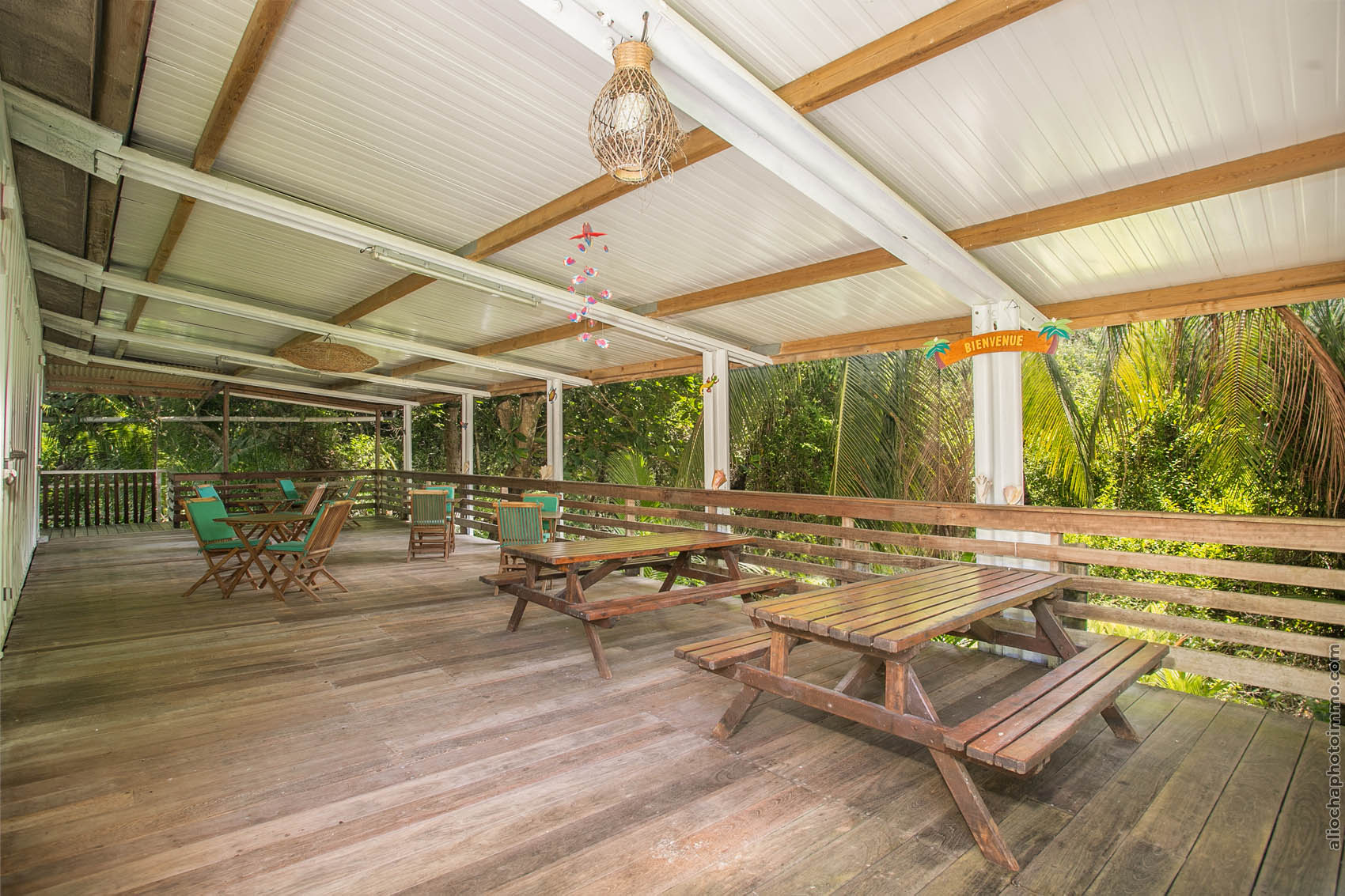 Location de salle ouverte sur l'extérieur en Martinque Sainte Luce - Véranda en bois couverte, équipée de tables de pique-nique et de chaises aux coussins verts, donnant sur une végétation tropicale luxuriante.